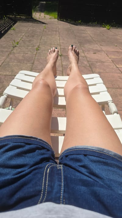 The image shows a person sitting in a white plastic lawn chair on a brick patio. The person is wearing denim shorts. The lower legs and feet are extended towards the left side of the frame. The toenails are painted with dark polish. The location is outdoors, with a glimpse of greenery and a fence in the background on the left. The brick patio is in the foreground.