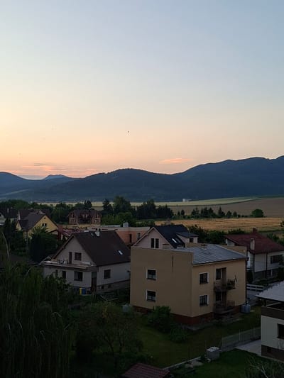 The image depicts a residential area in Ilava, Slovakia, during twilight. The foreground is dominated by the roofs of several houses, constructed with dark and light-colored tiles. One house in the mid-ground is a tan color with a metal roof and a small balcony. To the left, a large tree with drooping branches extends into the frame. In the background, a line of dark, tree-covered hills forms the horizon, with a patch of cultivated fields visible between the houses and the hills. The sky is clear and fades from a pale peach near the horizon to a pale blue at the top of the frame, with a few small birds silhouetted against it. The overall impression is a quiet, rural evening scene.