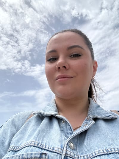The media file is a portrait of a young woman against a cloudy sky. She is wearing a light blue denim jacket with visible stitching and silver snaps. A delicate gold necklace is visible beneath the collar of her jacket. Her hair is dark and pulled back, with a few strands loose around her face. She has expressive dark eyes, lightly made-up eyebrows, and her lips are slightly parted, suggesting a relaxed or contemplative expression. A small hoop earring is visible in her left ear. The perspective is a low-angle shot, looking up at the woman. The background is dominated by a sky filled with white and grey clouds, indicating it is daytime and possibly overcast. There are no visible text elements in the image. The overall impression is a casual, candid moment captured outdoors. The location context provided is Foia L'om, Spain.