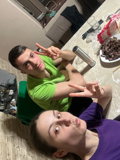 A selfie-style image depicts a man and a woman positioned around a table. The man, wearing a green t-shirt, is on the left, making a peace sign with his hand. The woman, in a purple t-shirt, is on the right and has her lips pursed. On the table, there are a plate of cooked meat, a metal thermos, a glass, a remote control, and packaging materials. The tablecloth has printed designs. In the background, there is a wall and furniture including a couch and decor items. The image is taken indoors. There is no explicit evidence indicating a location in Vinarovo, Bulgaria.