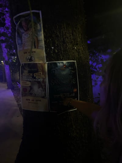 Three posters are attached to a tree trunk at night. The posters are for events happening at the Banana Beach in Cobano District, Costa Rica. The posters advertise a beach soccer tournament, a live show, and an event called "Tasty Santa Tere." The image shows a person reaching towards the posters, seemingly reading them. The scene is located in the Cobano District, Costa Rica, at latitude: 9.633, longitude: -85.158. The posters are lit with a faint blue light. The surrounding area is dimly lit, and the tree trunk is visible in the background.
