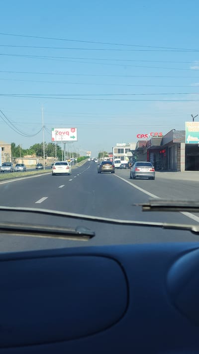 A wide-angle, eye-level shot shows a road in Ptghunk, Armenia, on a clear day. The asphalt road has white lane markings and is bordered by sidewalks and buildings. Several cars are driving on the road, including a white sedan in the foreground, a dark sedan in the middle lane, and other vehicles further ahead. Buildings with signs are visible on the right side of the road, including one with "CPS" written on it. A billboard with an arrow is seen on the left side of the road. The sky is blue with some faint clouds. The shot is taken from inside a vehicle, with the windshield wipers and dashboard visible in the foreground.