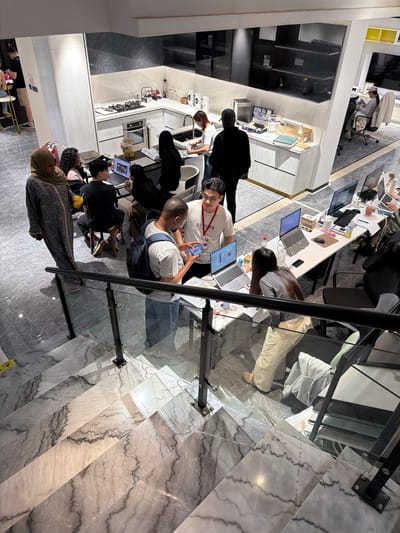 The image depicts an indoor scene, likely a modern office or co-working space, observed from a high vantage point, perhaps a mezzanine or upper floor. The location is specified as Chancheng District, China.
Several individuals are present, engaged in various activities. Some are working at desks equipped with computers and other office supplies. One person is looking at a phone. Some are interacting with each other.
The environment includes office furniture such as desks, chairs, and storage units. There's also what appears to be a kitchen area with appliances and counters. The floor is tiled with a patterned design. There are railings and glass partitions suggesting a multi-level structure. Artificial lighting illuminates the space.
The interaction and actions involve work related activities, looking at a phone and general office interactions.