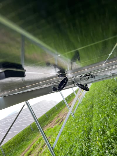 The image shows a close-up of solar panels, likely located outdoors in a rural area. The panels are arranged in a row, with visible electrical connections and mounting hardware. Green grass is visible in the background, indicating a natural environment. Although Grange Ville, Ireland, is mentioned, there is no definitive proof to verify this location.