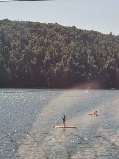 Two people are paddleboarding on a body of water. The person on the left is standing upright on a paddleboard, facing away from the camera. The person on the right is sitting on a paddleboard with another person. The water is sparkling with sunlight. In the background, there is a dense forest covering a hillside, and a clear sky above. There are also some faint horizontal lines visible in the foreground, possibly from power lines or a fence. The location is the city of Pavlovka, Russia.