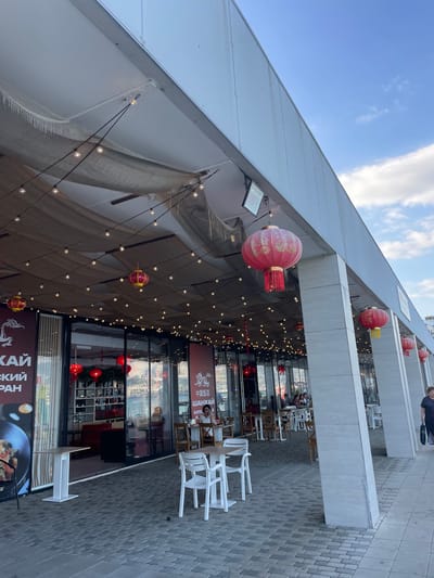 The image captures an outdoor seating area of a Chinese restaurant, possibly during daytime. The setting appears to be urban, with a paved walkway leading to the establishment. Overhead, a white roof structure extends outwards, adorned with strings of small, warm-toned lights and several large, red Chinese lanterns. The lanterns are decorated with intricate patterns.

The restaurant's interior is visible through large glass windows, revealing tables and chairs, as well as shelving units. Some of these tables are set up outside, with white plastic chairs and wooden chairs arranged around them. People are seated at some of the outdoor tables, engaged in conversation and dining.

The sky above is partly cloudy, suggesting a pleasant, possibly warm day. The architecture of the building is modern, with clean lines and a white facade. There is visible text on the building's signage. One sign clearly displays Chinese characters ("中国饭店" which translates to "Chinese Restaurant") alongside Russian text ("ШАНХАЙ" which translates to "Shanghai"). Another sign features the Russian phrase "КАЙСКИЙ ПРАН", also related to the restaurant's theme. The overall atmosphere seems relaxed and inviting, suggesting a leisurely dining experience.
