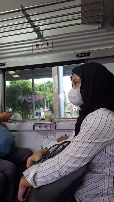 The image captures an interior view of a train carriage, likely in the city of Karawang, Indonesia, based on the contextual information. In the foreground, a person wearing a black hijab and a white and brown striped shirt is seated, looking out the window. They are also wearing a white face mask. Their arms are resting on a black bag. To the left, another person is partially visible, wearing a blue shirt and dark pants, holding a smartphone. Through the window, lush green trees and some buildings are visible, suggesting a semi-urban or suburban landscape. On the train wall, there are emergency instructions with text in Indonesian and English. Electrical outlets are also visible on the wall below the window.