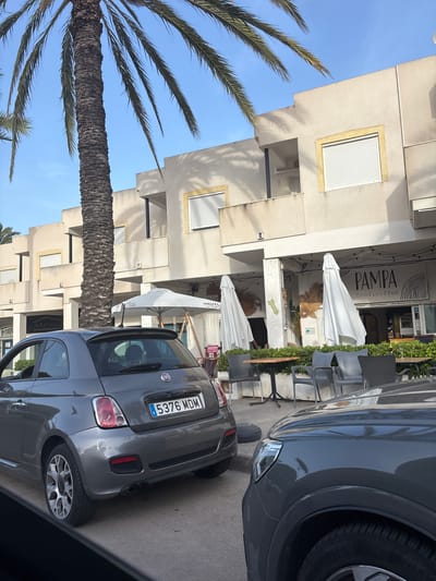 A grey Fiat 500 automobile is parked on a paved road in front of a building with a palm tree to its left. The building is multi-story with light-colored stucco walls and white-shuttered windows. An awning extends over the ground-floor storefronts. One storefront is labeled "PAMPA food & coffee." Outdoor tables and chairs are arranged in front of the storefronts. The car's license plate reads "E 5376 MDM". The sky is clear and blue. The scene is located in Urbanización Cabo Roig, Spain.