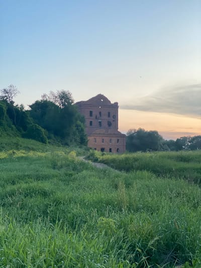 The image depicts an abandoned brick building situated on a grassy hill in Yuryatino, Russia. The building, which appears to be a former mill, is in a state of disrepair, with broken windows and sections of its roof missing. The surrounding landscape is characterized by lush green grass and trees, suggesting a rural or natural setting. The sky above is clear with a hint of sunset colors on the horizon. There are no people or animals visible in the scene.