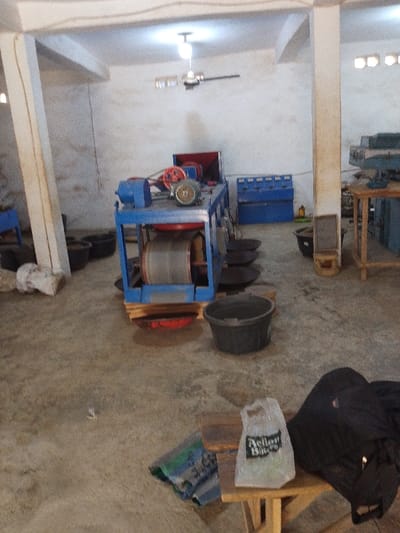 The image depicts the interior of an industrial workshop or processing facility located in Olomoda, Nigeria. The concrete floor is visible, and the walls are light-colored. Exposed concrete pillars support the ceiling, where a single incandescent bulb illuminates a ceiling fan.
The central subject is a blue industrial machine, likely a trommel or processing unit, positioned on wooden pallets. It features a red input hopper at the top, a blue electric motor, a grey/silver electric motor with red pulleys and drive belts, and a large cylindrical mesh drum. Below the drum, a red collection pan is situated on the floor.
Around the central machine, several dark-colored plastic buckets are present, some appearing to contain material. To the right of the machine, approximately five dark, shallow, circular pans are arranged on the floor. In the background, a blue multi-compartment cabinet is visible. Further right, a wooden workbench or table holds additional equipment.
In the foreground, a small wooden stool supports a white plastic shopping bag with the "Action Bitters" logo and a folded black jacket. A green and blue mat or tarp lies on the floor near the stool. Near a left-side pillar, a white sack rests on the floor. No individuals are present in the scene, and no active processes are underway.