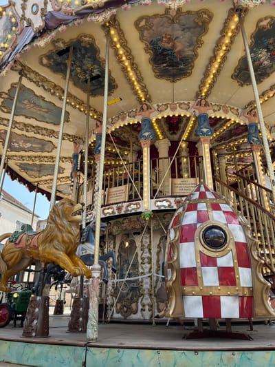 This is a close-up, eye-level, outdoor shot of a carousel, likely a popular attraction in Rambouillet, France. The carousel is ornate, with a pale yellow ceiling adorned with paintings of cherubs and floral motifs, framed by decorative golden scrollwork. Rows of small, yellow lights line the ceiling's edge, illuminating the scene.
The carousel features several animal figures, most notably a striking golden lion prominently positioned on the left, with its mouth open and teeth bared. Behind the lion, dark horses are visible, and further back, other carousel figures and decorative elements can be seen. The structure includes ornate columns topped with figures of mermaids or sirens. A red and white checkered, rocket-shaped structure is also a prominent element. In the background, a hint of urban architecture, including buildings and a bright blue sky, suggests the carousel is situated in a town. To the far left, a vintage green tractor with red wheels is partially visible. Several signs are attached to the carousel structure, some with French text, indicating rules or information for riders. The overall impression is one of a vibrant, classic amusement ride.