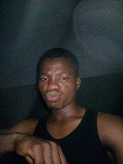 In a dimly lit room in Eleweran, Nigeria, a young man with dark skin and short hair is seen taking a selfie. He is wearing a black tank top and has his lips puckered as if to give a kiss. His left arm is resting on his lap, and his fingers are slightly curled. The background is a dark, textured wall. The lighting is low, casting shadows on his face and the surrounding environment.