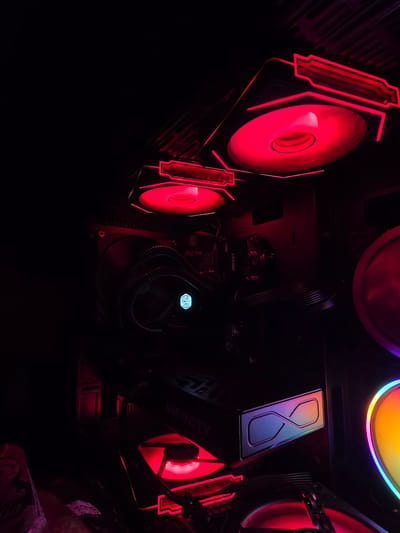 In the dark interior of a computer case, a red LED light illuminates components. Two fans with red lighting are visible at the top of the case. A Cooler Master liquid cooler with a blue logo is positioned on the left. Below it, a graphics card with the word "INFINITY" and an infinity symbol is visible. Red LED strips line the bottom of the case, illuminating another fan and parts of the graphics card. The scene is enclosed within the computer case, with no external environment visible. The specific location in Aleksandrovo, Bulgaria is not discernible from the image.