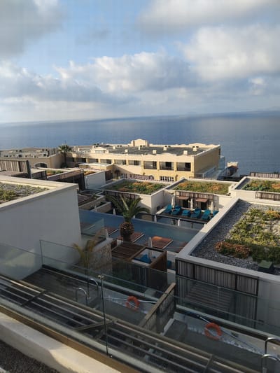 This is an outdoor, aerial view of a resort in Pyli, Greece, taken during daylight hours. The resort complex is located alongside the sea, with blue water stretching to the horizon under a sky with scattered white clouds. The buildings have a modern, layered architectural style, with light-colored facades. Some of the roofs are designed as green spaces, featuring vegetation and landscaping. There is a swimming pool area with blue sun loungers and other seating arrangements. A glass-paneled balcony is in the foreground, with metal railings and an orange life preserver attached to the wall. There are no people immediately visible in the scene.
