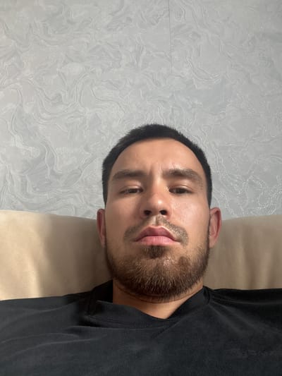 This is a close-up, head-and-shoulders photograph of a young man. He is lying down, with his head turned towards the camera. His eyes are open and he is looking directly at the viewer. He has short, dark hair, a short beard, and light brown eyes. He is wearing a black t-shirt. The background consists of a light-colored, patterned wall and a beige surface, possibly a bed or couch. The lighting suggests an indoor setting, likely during the daytime. The man's expression appears neutral or perhaps slightly pensive. There is no visible text in the image. The provided location context is Достық ауылдық округі, Kazakhstan.