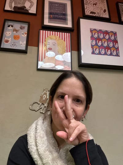 A woman angles her head towards the camera, holding up two fingers in a peace sign. She's wearing a black shirt with red trim and a fuzzy, cream-colored scarf. Behind her is a wall, divided into two colors, with a display of framed artwork including illustrations of cats, portraits, and text in Italian. Also visible is a decorative, sparkly reindeer head mounted on the wall.