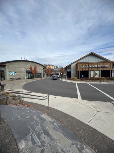 A tilted outdoor shot showcases a street view with buildings on either side. "American Eagle Outfitters" signage is visible on one building. A road with a white stripe runs between the buildings, and a black car is seen driving on it. Steps and railings occupy the lower right corner, appearing worn. The sky is cloudy and blue.