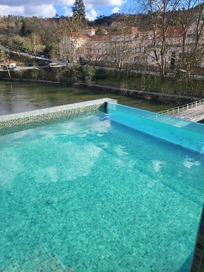 The image displays an elevated outdoor infinity pool, tiled with small turquoise mosaic squares. The pool water reflects patches of white clouds and blue sky. Immediately beyond the pool's infinity edge, a greenish-brown river flows horizontally across the mid-ground. Along the river's far bank, a narrow paved path or road is visible, accompanied by a line of mature deciduous trees largely devoid of leaves. Further back, situated on the riverbank, are several multi-story buildings featuring light-colored facades and distinctive reddish-orange tiled roofs, typical of traditional Portuguese architecture. These structures are nestled amidst additional bare trees. To the far right, a white pedestrian bridge with railings extends along or crosses a section of the river. The background consists of densely wooded hills under a sky with scattered white clouds. No individuals or specific actions are depicted. The scene is located in São Pedro do Sul, Portugal.