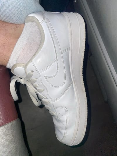 A person is wearing a white Nike Air Force 1 sneaker with white laces and a gray sock. The shoe has a thick white sole. The person is wearing jeans on one leg and sitting on a maroon surface. A gray object is slightly visible in the background. The lighting is subdued.