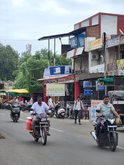 The image depicts a street scene in Datala, India, under an overcast sky. In the foreground, two men are riding motorcycles. The man on the left is riding a black scooter with a red container attached to the side. He is wearing a white shirt with blue stripes and dark pants. The man on the right is riding a black motorcycle, wearing a light blue polo shirt and dark pants. He is carrying a bag on the front of his motorcycle. In the background, a busy street is lined with shops and buildings. A cell tower is visible in the distance. Several other motorcycles and a fruit stall with a yellow awning are also present. The overall environment appears to be an urban setting with moderate pedestrian and vehicular activity.