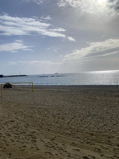 An aerial view shows a beach in Garrucha, Spain, with the Mediterranean Sea in the background. The sky is partly cloudy, with sunlight glinting off the water. In the foreground, a sandy beach is visible, with a volleyball net set up. A yellow ATV is parked to the left of the net. In the distance, several cargo ships are anchored in the sea, and a pier extends into the water. The waves are gently lapping at the shore, and there are a few buoys scattered in the water.