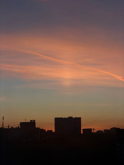 The image displays an urban twilight scene in the city of Shul'gino, Russia, observed during either sunrise or sunset. The sky occupies approximately three-quarters of the frame, exhibiting a distinct color gradient: dark purplish-blue at the zenith, transitioning to a prominent orange-pink hue across the mid-sky, and fading to a lighter orange-yellow near the horizon. Thin, wispy cloud formations are visible within the orange-pink strata. A single, linear condensation trail extends diagonally from the upper-left to the mid-right, appearing brighter against the surrounding sky.

The lower quarter of the image features the silhouette of an urban skyline. Multiple multi-story buildings are present as dark, indistinct forms. A tall, rectangular building is visible on the far left, while a large, blocky structure dominates the central skyline. Several smaller, less defined buildings are scattered across the horizon line to the right. Thin, dark lines, consistent with power or communication cables, traverse horizontally across the mid-ground, connecting between unseen points and visible building structures. A tripod-like antenna or mast is faintly discernible on a rooftop towards the left side of the frame. No individuals, specific actions, or direct interactions are identifiable within the scene.