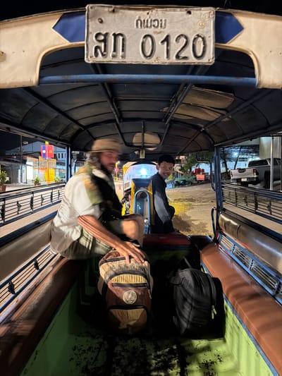 The image shows the interior of a tuk-tuk at night in Thakhek, Laos. A man wearing a hat and a white shirt is seated on the left side of the tuk-tuk, with his arm resting on a brown and tan backpack. On the right side, a young man in a dark shirt is seated and looking at the camera with a smile. Two backpacks are visible in the tuk-tuk, one brown and tan, the other black. The license plate on the front of the tuk-tuk reads "สท 0120" and has some Thai or Laotian script above it. In the background, buildings and streetlights are visible, along with a parked pickup truck. A flag with red and yellow colors, possibly the flag of Laos, is also visible. The overall environment suggests a street scene in a Laotian city at night.