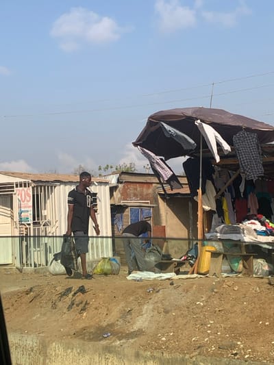 The image depicts an informal street market scene in Abobo, Nigeria, under a clear, bright blue sky with sparse clouds. The foreground consists of uneven, dry, light brown earth with scattered small debris, bordered by a concrete or stone curb in the lower frame. Overhead, thin utility wires span the scene.
On the left, a male with dark skin, wearing a black polo shirt, dark shorts, and dark slide sandals, walks towards the right. He carries one black plastic bag in each hand and has a white earpiece in his right ear. A light-colored, possibly white or light green, sack rests on the ground near his left foot.
In the mid-ground to the right, a second male figure, also with dark skin, is bent over, facing away from the viewer. He wears a dark long-sleeved shirt and light gray sweatpants, and appears to be rummaging through a large, light-colored bag or a pile of items on the ground next to a small wooden table. Behind him, an informal market stall is constructed with a dark brown or black umbrella serving as a primary canopy, supported by wooden poles. Various shirts, including checkered patterns, are hung from the umbrella's frame and the supporting structure. Piles of additional clothing items are displayed on surfaces beneath the canopy. A yellow/green sack is visible on the ground near the market stall.
The background features several buildings of varying construction. To the left, a structure with a white corrugated metal facade displays a red sign reading "POS SERVICES" and has barred windows. Adjacent to it, a more weathered building with a corrugated metal roof, exposed brick or plaster, and a blue-framed window is visible. Clothes are draped on a line or rail in front of this building. Further structures with corrugated metal roofs are visible in the distant background.