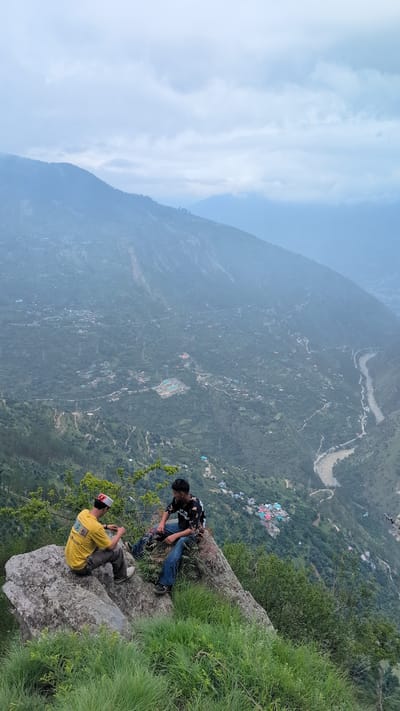 Two young men are seated on a large rock on a steep, grassy incline overlooking a valley. The man on the left wears a yellow t-shirt and a red and white patterned hat. He is facing the man on the right and appears to be talking to him. The man on the right wears a black t-shirt with a white graphic and blue jeans. He is looking down at the valley, with his legs dangling over the edge of the rock. The valley below is densely covered in trees and scattered with small buildings and a winding river. The distant mountains are shrouded in mist, creating a hazy effect. The overall scene is one of serene contemplation in a vast, natural landscape. The specific location is Cheelaage, India, a region known for its mountainous terrain and scenic beauty.