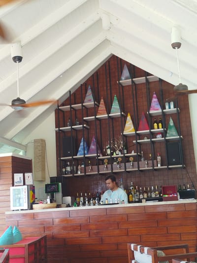The image shows a bar area at a resort in Gulhi Falhu, Maldives. A bartender in a light blue shirt is standing behind the counter, arranging bottles. The bar is made of dark wood and is stocked with various bottles of alcohol and glasses. Behind the bar, shelves are decorated with colorful, triangular art pieces and more bottles. A ceiling fan is visible above, and the ceiling is sloped and made of white wood planks. The overall impression is of a well-appointed bar in a tropical setting.