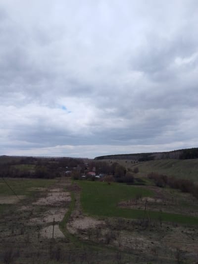 This wide-angle landscape image captures a tranquil rural scene under an overcast sky, likely in Znamenka, Russia. The sky dominates the upper half of the frame, filled with dense, light grey clouds, with a tiny patch of brighter blue visible towards the upper left.

The foreground consists of rugged, uneven ground covered in dry, sparse vegetation and patches of grass, suggesting either early spring or late autumn. A narrow, unpaved track or dirt road winds through this area, curving towards the middle distance. Several utility poles with wires are visible along this path, indicating basic infrastructure.

In the middle ground, a small cluster of houses or buildings, characteristic of a village or hamlet, is nestled amongst trees and green fields. Some of these fields are noticeably greener and more cultivated, contrasting with the drier patches. The trees surrounding the buildings appear to be deciduous, with some bare branches, further hinting at a non-summer season.

Beyond the village, the landscape rises into rolling hills, covered with a mix of scrubland and a dense forest of dark trees forming a distinct horizon line in the far background. The overall impression is one of quiet solitude.

There are no people or specific activities visible in the image. The time of day appears to be daytime, but the heavy cloud cover gives a subdued and soft lighting, suggesting an overcast day rather than bright sunshine. No visible text is present in the image.