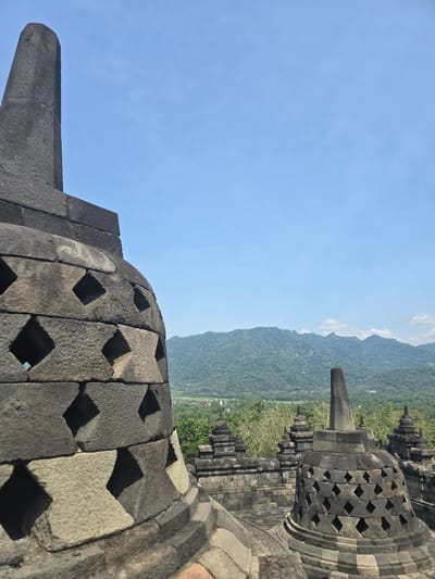 The image captures a partial view of Borobudur, a 9th-century Mahayana Buddhist temple located in Desa Borobudur, Indonesia. The foreground features the distinctive perforated stupas, constructed from dark-colored stone blocks. The geometric shapes of the perforations form diamond-shaped openings. The perspective suggests the photograph was taken from within the temple complex. In the background, a mountain range is visible under a clear blue sky with wisps of clouds. The landscape between the temple and the mountains appears lush with vegetation.