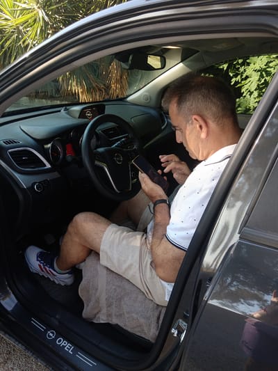 In the Municipal Unit of Vocha, Greece, a man is sitting in the driver's seat of a car, looking at his mobile phone. The car is gray, and the interior is dark. The man is wearing a white polo shirt with a striped collar and beige shorts. He has short, graying hair and is wearing a watch on his left wrist. His right foot is resting on a car mat with an "Opel" logo. Outside the car, there are palm trees and green foliage visible. The sun is shining, casting shadows and creating bright highlights on the car's surface.