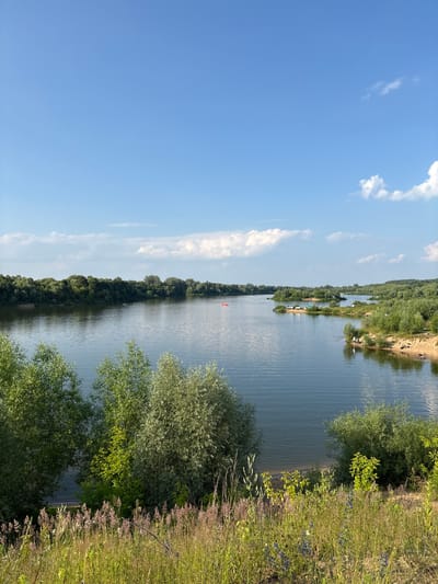 The image depicts a wide, calm river under a bright blue sky with scattered white clouds. The riverbanks are lined with dense green trees, and in the foreground, there is a grassy area with wildflowers. On the far side of the river, a line of trees forms a natural border. In the middle of the river, a small red boat can be seen. Further along the right bank, there are sandy areas and patches of vegetation, with a few small groups of people visible near the water's edge. The scene is characteristic of a natural landscape within or near the city of Lan'shino, Russia, likely a popular spot for recreation during warm weather.