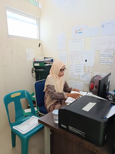 In Kuta Pungwa, Indonesia, a woman wearing glasses and a hijab is seated at a desk in a room that appears to be an office. She is looking down at documents on the desk and appears to be working. The desk is cluttered with papers, a computer monitor, a fan, a printer, and a glass of water. Behind her, the walls are covered with various documents and papers, some of which are pinned or taped up. There is a window on the left side of the room, and a stack of folders and binders is visible against the wall. A bright blue chair is situated to the left of the desk. The overall impression is that of a busy, functional workspace.