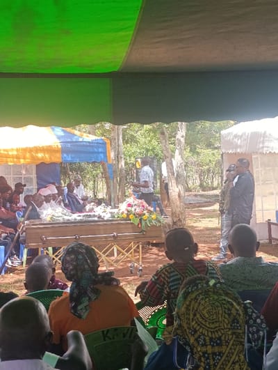 An outdoor funeral service is underway in Muthetheni ward, Kenya, under various temporary shelters. In the foreground, numerous individuals are seated on green plastic chairs, viewed primarily from behind. A woman in an orange top and a multicolored headscarf is prominently centered, with "KAVI" visible on her chair. Attendees hold papers, likely programs.
At the center of the scene, a light brown wooden coffin rests on a gold-colored collapsible gurney. The coffin is adorned with an arrangement of pink, yellow, orange, and white flowers. Behind the coffin, a group of individuals, including some in white garments, are seated.
To the right of the coffin, a man in a light-colored patterned shirt stands, holding a clear bottle. Further right, two men stand: one in a black jacket and sunglasses holds a microphone, next to another man in jeans and a light shirt.
The area is illuminated by bright sunlight, with the ground composed of dirt. Several sparsely foliated trees are visible in the background, defining the natural, open environment. Overhead, the main canopy covering the foreground is green and dark-colored. A smaller blue and yellow striped tent is situated to the left, and a white tent is visible on the far right.