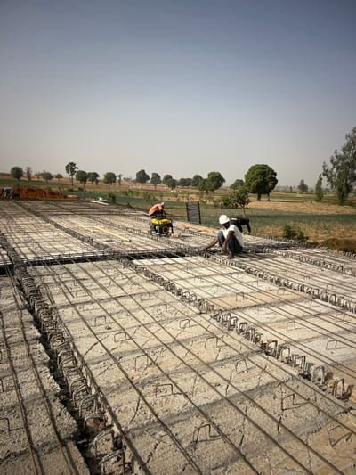 The image depicts an active construction site, likely for a large concrete structure such as a bridge deck, road, or an extensive floor slab. The perspective is slightly elevated, looking down and along the length of the structure.
In the foreground and midground, a dense network of steel reinforcement bars (rebar) is laid out. This rebar grid consists of numerous longitudinal bars running parallel to each other, interconnected by regularly spaced stirrups or ties that form rectangular cages. This intricate rebar assembly is positioned on top of an existing, somewhat rough-textured concrete surface, which forms the base layer for the new pour. The concrete base shows patterns or marks from its previous casting, indicating it might be a pre-existing part of the structure or a blinding layer.
Several workers are engaged in activities on this rebar structure. In the midground, one worker, wearing a white hard hat, a blue shirt, and dark trousers, is kneeling on the rebar, seemingly inspecting, tying, or adjusting the steel bars. Another worker in a dark shirt is partially visible behind him. Further down the structure, towards the left, another worker in an orange shirt and dark trousers is standing next to a yellow portable machine, which appears to be a small engine-driven device, possibly a concrete vibrator or a generator. Other workers are vaguely visible in the distance, suggesting a larger team.
The background reveals a rural or semi-rural landscape under a clear, bright sky. Fields, possibly agricultural, with varying shades of brown and green, stretch into the distance. A line of green-leafed trees, some isolated and some forming small clusters, dots the landscape, suggesting boundaries or distant vegetation. The lighting indicates bright daylight, possibly midday, with a uniform, slightly hazy quality to the sky.
The overall scene conveys an impression of ongoing, large-scale civil engineering work, characterized by manual labor and the precise arrangement of reinforcing steel in preparation for a concrete pour. There is no visible text in the image.