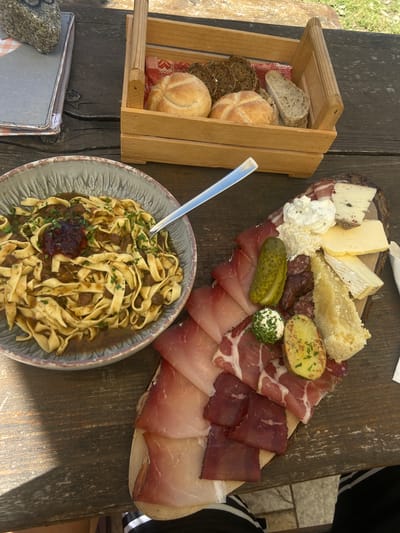 A rustic wooden table is set with a selection of food and drinks, suggesting an outdoor meal or gathering. In the foreground, a bowl of pasta with a dark sauce and finely chopped parsley is prominently featured, with a fork resting in it. To the left, a wooden crate holds various types of bread, including sliced rustic loaves and round rolls, nestled on a red and white checkered cloth.

Dominating the upper right side of the image is a heart-shaped wooden board laden with a charcuterie and cheese platter. It showcases an assortment of cured meats, several types of cheese, a dollop of creamy white spread, a large pickle, a small ball of cheese covered in herbs, and a halved baked potato sprinkled with herbs. The entire spread is artfully arranged on the textured, rustic surface of the board. The scene is set on a wooden table, with hints of greenery and a paved area visible in the background, suggesting an outdoor dining setting, possibly in a garden or patio area. The lighting suggests it is daytime, with sunlight casting shadows on the table, indicating a sunny day. The presence of a person's arm and leg in black and white striped clothing on the far right hints at a person being present and enjoying the meal, but they are not the main subject. The overall impression is one of a hearty and enjoyable meal served in a casual, outdoor environment.