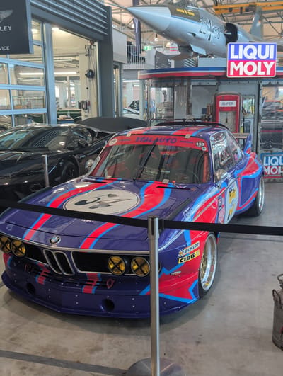 This media file captures a vintage BMW race car, likely displayed in a museum or exhibition setting in Böblingen, Germany. The car is predominantly purple with red and blue racing stripes, featuring prominent "LIQUI MOLY" and "DUNLOP" branding. A large white circle with a black emblem is visible on the side of the car. In the background, a vintage airplane, possibly a F-104 Starfighter given the German context, hangs suspended. A classic red gas pump with the word "SUPER" is also visible, adding to the automotive theme. The overall scene suggests a car show, museum, or event celebrating automotive and aviation history. The lighting appears to be artificial, consistent with an indoor exhibition space. The image is oriented vertically. Visible text includes "LIQUI MOLY", "SUPER", "BENNWAGEN", "LEGENDS", "SOHNGEN DESIGN.COM", "DUNLOP", and "CIBIE".