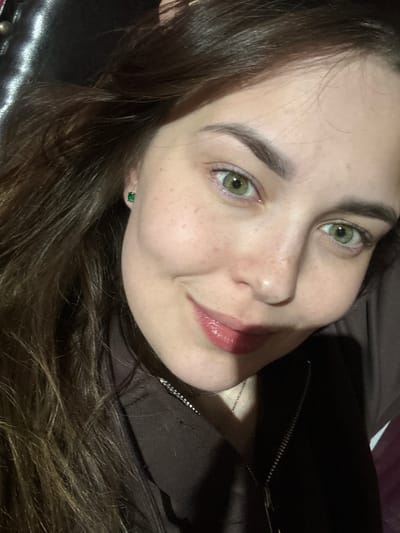 A young woman with light skin, freckles, and green eyes poses for a selfie. She has long, dark brown hair that is slightly messy. She is wearing a dark brown zip-up hoodie and a small, emerald green stud earring in her left ear. Her lips are slightly parted and appear to be wearing lip gloss. The background is dark and out of focus, with some shiny surface visible on the left. The image was taken indoors. There is no information available regarding the location in the city of Zhebrei, Russia.