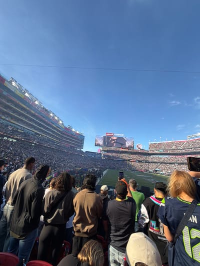 A stadium packed with people, possibly for a Super Bowl event, features a large screen displaying an image of a person. A green football field stretches out below the screen. A clear blue sky is visible. Spectators fill the stands, with some taking pictures. The image is slightly tilted. Levi's Stadium logo visible on the large screen.
