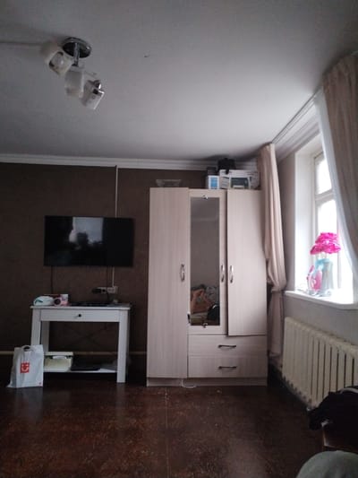 The image shows an interior scene, likely a bedroom or living area, with a dark brown wall on the left and a light-colored wall on the right. A flat-screen television is mounted on the left wall. Below the TV, a white table with a drawer holds various items including a pink speaker, a package, and a remote control. To the right of the TV, a light-colored wardrobe with mirrored doors is visible. On top of the wardrobe, several boxes are stacked. A window with light-colored curtains is present on the right side of the image, with a radiator beneath it. A pink floral arrangement sits on the windowsill. The floor is made of dark wood. A ceiling light fixture with three adjustable lamps is visible at the top of the image. There is no visible indication of the city of Muhametjan Tüymebaev, Kazakhstan.