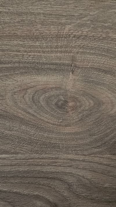 The image is a close-up, vertical shot of a weathered, greyish-brown wooden surface. The grain of the wood is prominent, featuring swirling patterns around a distinct knot or growth ring in the center. Fine lines and subtle cracks are visible throughout, indicating age and exposure. The lighting is even, highlighting the texture without harsh shadows. There are no people, objects, or any environmental context visible in the image; it is solely a textural study of wood. The location in the city of Shali, Russia, is not discernible from the image itself.