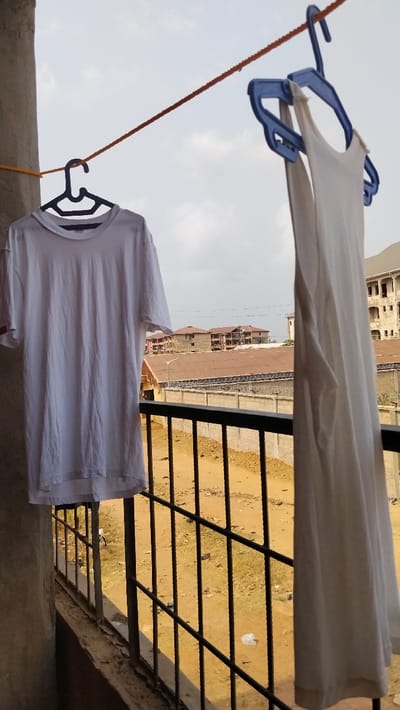 The scene depicts a view from a balcony in Isieke, Nigeria. An orange rope, serving as a clothesline, stretches diagonally across the upper portion of the frame. A white short-sleeved t-shirt hangs on a black plastic hanger from the left side of the rope. To its right, a white sleeveless tank top is suspended from the rope on a blue plastic hanger with a double-loop design. A light-colored concrete pillar is partially visible on the far left. Below these items, a black metal balcony railing extends horizontally across the lower section of the image. Beyond the railing, the ground consists of exposed reddish-brown dirt with scattered litter, including a crumpled white plastic item. A concrete wall topped with coiled barbed wire is visible horizontally in the midground, behind which are structures with corrugated metal roofs and a streetlamp. In the background, multiple multi-story buildings, some in various stages of construction, are discernible against a partially cloudy sky.