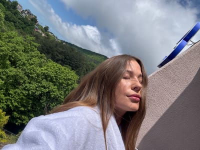 A woman is lying on a balcony overlooking a cityscape near Sosnovy Bor, Russia. She wears a white bathrobe. Behind her is a textured wall and, in the distance, a view of a hill covered in greenery and residential buildings. The sky is visible with some clouds.