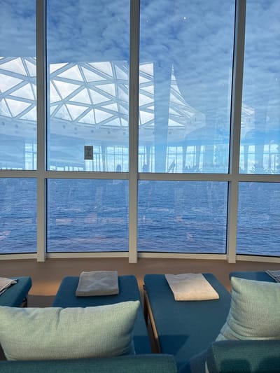 The scene depicts an interior lounge space with panoramic windows offering a direct view of expansive blue ocean waters. The location is specified as the waters off Long Island, Bahamas.
In the foreground, several vacant lounge chairs are arranged, upholstered in a teal or blue-green fabric. Each chair features a matching pillow, and at least two chairs have neatly folded light-colored towels placed on them. One towel clearly displays a white 'X' motif. The floor is a light brown, featureless surface.
The midground is dominated by large, white-framed windows spanning horizontally and vertically, segmenting the view. Through these windows, the deep blue water of the open sea is visible, showing subtle wave patterns. Above the water line, a bright blue sky with scattered white clouds is discernible. Reflections within the glass reveal a large, white, geometric architectural structure with a triangular grid pattern, likely part of the vessel or building from which the image is taken. Faint, indistinct human silhouettes are vaguely visible in the reflections or through the glass, suggesting other individuals in the vicinity. A small, dark rectangular sign is adhered to one of the central window panes.
The background consists entirely of the open ocean under a partly cloudy sky, extending to the horizon. No specific landforms or other vessels are visible. There are no individuals actively engaged in any discernible actions or interactions within the immediate frame.
