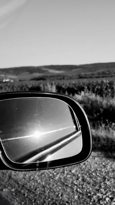 In Piatra Albă, Moldova, a black and white photograph captures a close-up view of a car's side mirror. The mirror reflects a bright sun, creating a lens flare effect, and a portion of what appears to be a road or grassy area. The background is out of focus, showing rolling hills covered in vegetation under a clear sky. The car's side mirror frame is visible, along with some scratches on the mirror's surface. The foreground shows a textured surface, possibly gravel or pavement.