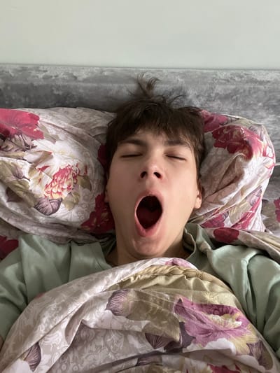 A single individual is observed lying in a bed in a room in Zainsk, Russia. The person, with short, dark brown hair, is captured in the act of yawning; their eyes are closed, and their mouth is wide open. The upper torso is covered by a floral-patterned duvet featuring red, pink, green, and cream hues, matching the pillow beneath their head. A light green long-sleeved garment is visible on their body. Behind the pillow, a grey, textured headboard is present, above which a plain light green wall forms the background.