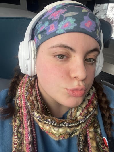 An individual is depicted in a close-up, self-portrait photograph. The subject, a young adult with fair skin, brown hair styled in two braids, and hazel eyes, looks directly forward with pursed lips. A small nose ring is present in the left nostril. They wear a dark teal head wrap patterned with pink, purple, light blue, and yellow flowers, positioned under white over-ear headphones. The visible earcup of the headphones displays "M/F" and "ON/OFF" labels. A multi-textured, multi-colored scarf, featuring shades of pink, brown, yellow, green, and purple, is wrapped around the subject's neck, worn over a blue long-sleeved garment. The background indicates an indoor travel environment, likely within a vehicle such as a train or bus, with a teal upholstered seat visible on the left and a blurred window view on the right suggesting a bright, possibly snow-covered, outdoor scene. No specific identifiable landmarks or signs within the image definitively confirm the location as Darien, United States of America; the context is limited to an indoor travel setting.