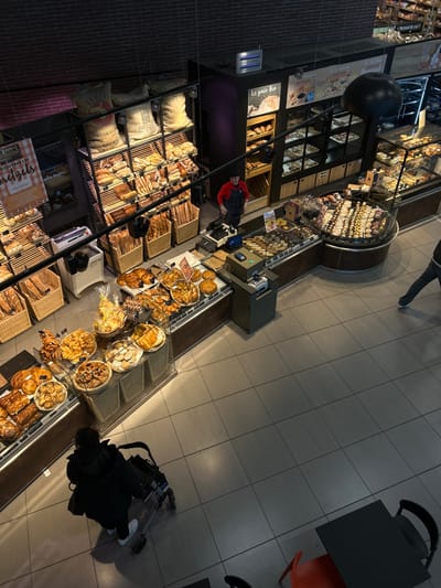 This image depicts the interior of a bakery section within a commercial establishment in Eschau, France, viewed from an elevated perspective. The scene is dominated by extensive displays of baked goods.
On the left, a multi-tiered shelving unit holds various breads and baguettes in wicker baskets. Above these shelves, several large burlap sacks are stacked. A vertical banner partially visible on the far left advertises "Découvrez les Bretzels." In front of these shelves, glass display cases present an assortment of pastries, including croissants and various sweet and savory items.
In the center-right, a long counter with additional glass display cases showcases a wide array of pastries, cakes, and tarts. An L-shaped section of this counter features a circular display unit filled with numerous donuts and small pastries. Behind this counter, an individual wearing a red shirt and a dark apron, presumably an employee, stands facing the displays. A cash register and payment terminal are visible on the counter. Above the counter, an insect light trap emits a blue glow. A sign reading "Le pain bio" (Organic bread) is visible on the wall behind the counter.
The floor is composed of large, dark gray tiles. In the bottom left, a person, viewed from behind, wearing a dark coat and pushing a black-framed shopping cart or stroller, is observing the pastry displays. On the far right, the lower legs and feet of another individual, wearing dark pants and white sneakers, are visible as they walk away. The far bottom right corner reveals a dark table with an orange chair and a black chair, indicating a small seating area. The ceiling and upper walls appear dark, with visible support structures and lighting fixtures.